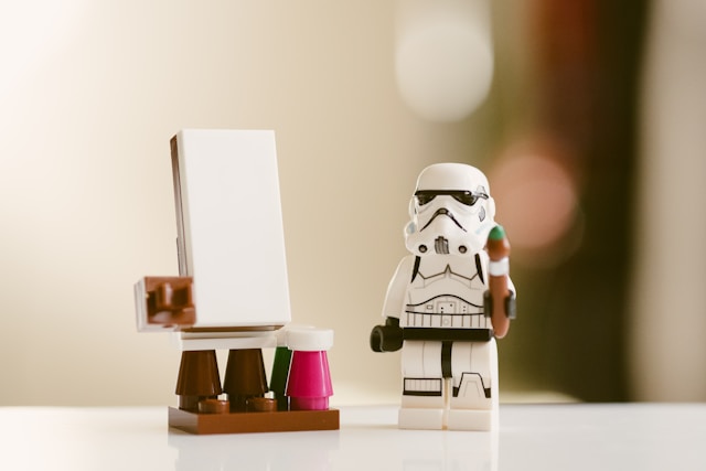 В центре изображена фигурка штурмовика из LEGO.
                  Рядом с ним находится мольберт с небольшим белым холстом. 
                  Возле мольберта — два маленьких контейнера, напоминающих баночки с краской.