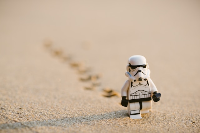 в центре внимания находится маленькая фигурка штурмовика из LEGO. 
                  Штурмовик идет по песчаной поверхности, оставляя за собой следы. 
                  Он изображен на переднем плане. На заднем плане видно размытую линию следов, 
                  которые исчезают вдалеке, акцентируя внимание на движении фигурки вперед.